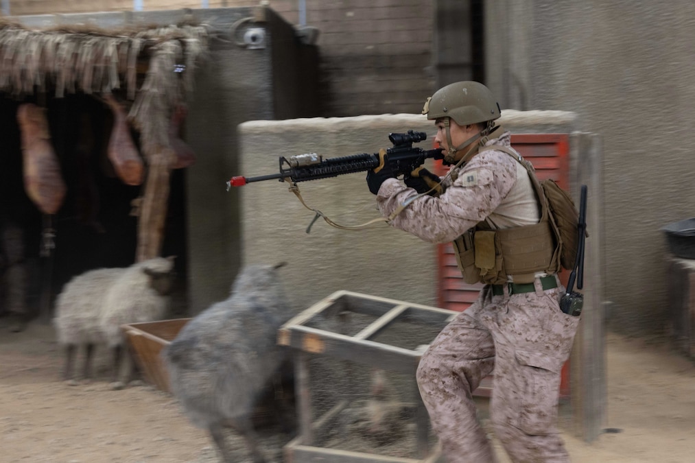 A Marine runs with a weapon with concrete structures with fake sheep and chickens nearby.