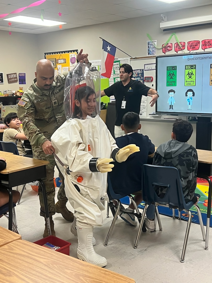 A student tries on a Biosafety Level 4 Positive Pressure Suit with assistance from Capt. Jesus Silvas, a microbiologist with the U.S. Army Reserve Medical Command assigned to the 59th Medical Wing’s Office of Research and Technology Applications, Aug. 7, 2025, at Mendez Elementary School in San Marcos, Texas, as Dr. Israel Guerrero, a virologist with Texas Biomedical Research Institute, describes laboratory safety levels.