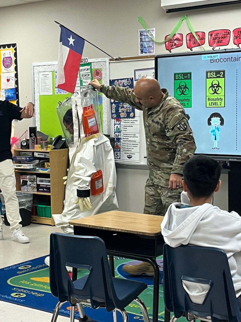 Capt. Jesus Silvas, a microbiologist with the U.S. Army Reserve Medical Command assigned to the 59th Medical Wing’s Office of Research and Technology Applications, shows students a Biosafety Level 4 Positive Pressure Suit Aug. 7, 2025, at Mendez Elementary School in San Marcos, Texas, as Dr. Israel Guerrero, a virologist with Texas Biomedical Research Institute, explains its use.