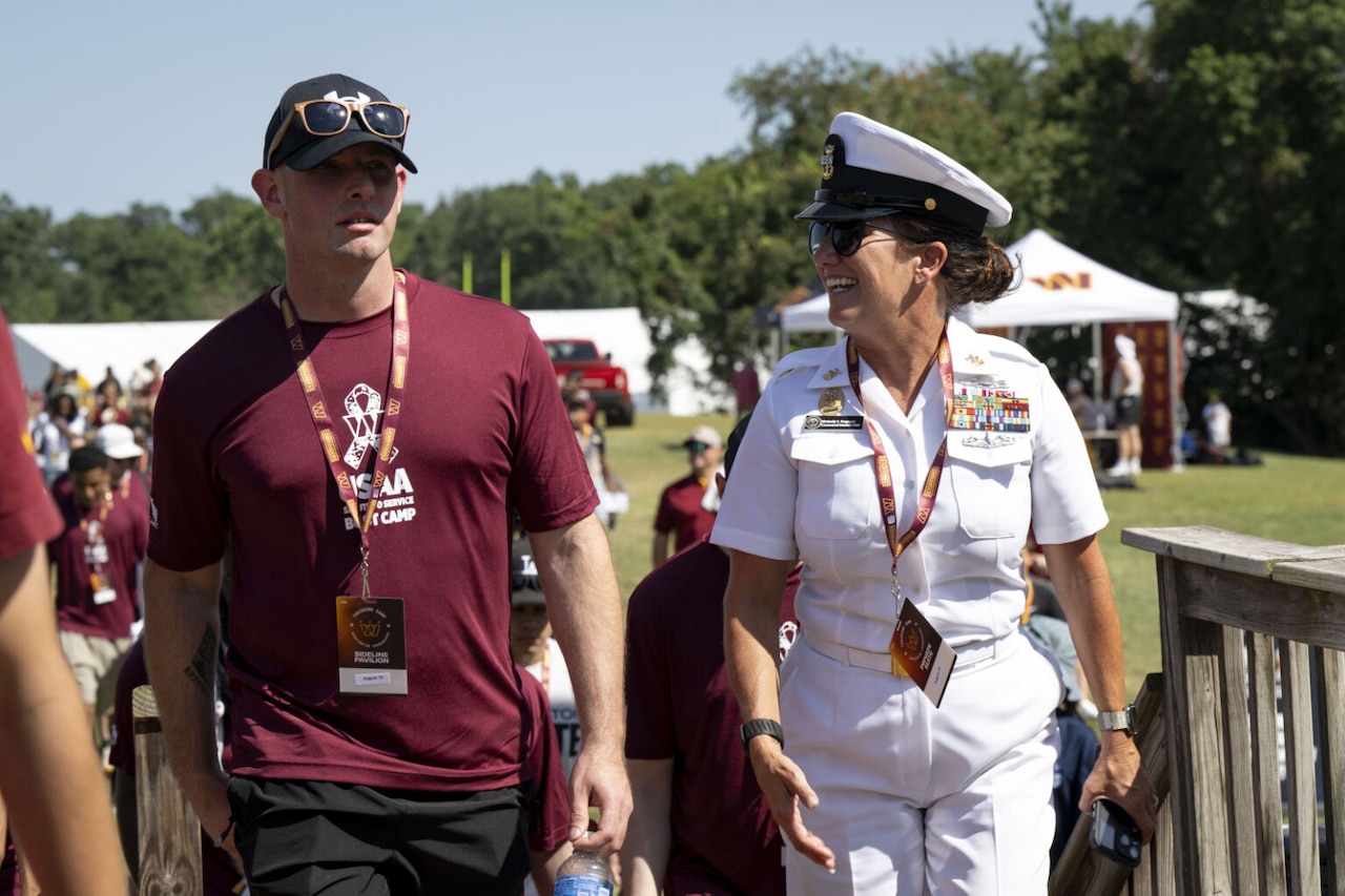 A man in a ball cap and T-shirt is walking outdoors next to a smiling woman in a military dress uniform decorated with ribbons and metallic devices.