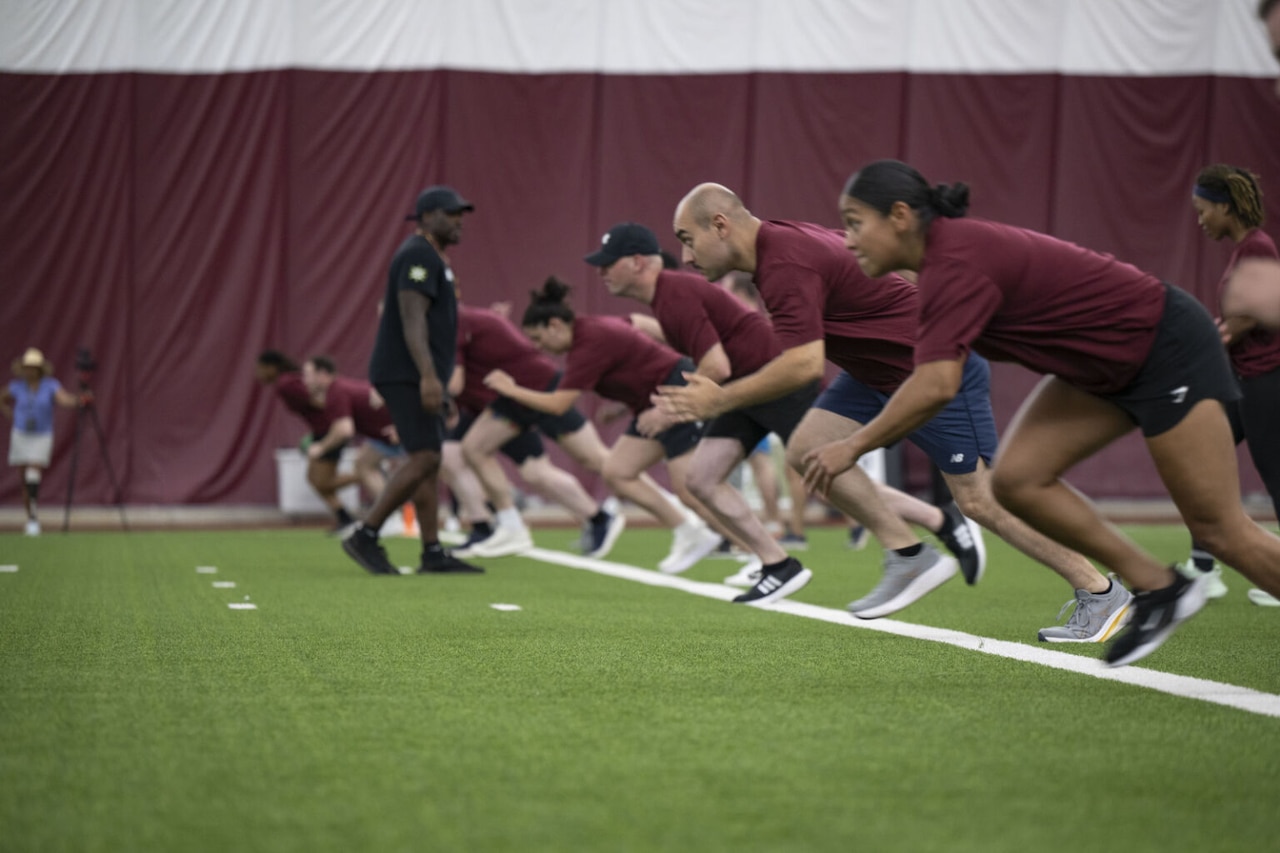 A group of approximately 10 men and women in athletic shorts and T-shirts are sprinting down a football field.