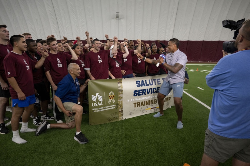 A group of men and women in athletic attire are standing on a football field, smiling and raising their hands while being spoken listening to a man holding up a microphone.