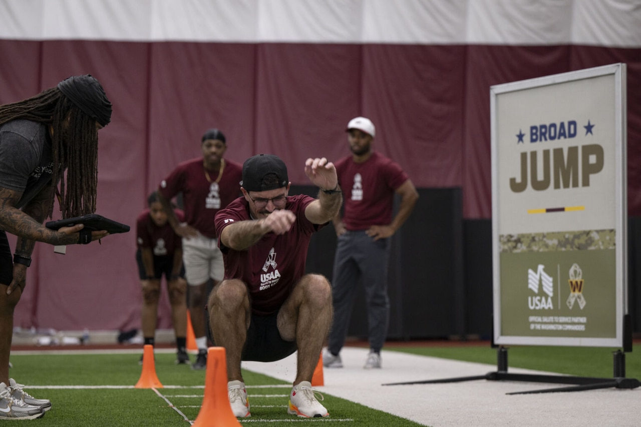 A man in athletic attire lands on a green football field after completing a broad jump. A person is standing nearby holding an electronic devise and observing the man’s distance.