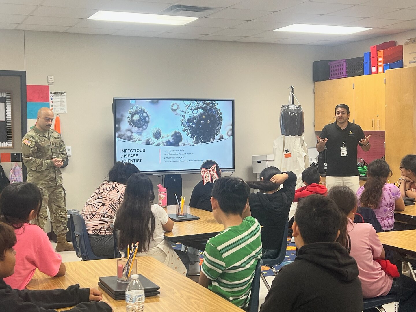 Capt. Jesus Silvas, a microbiologist with the U.S. Army Reserve Medical Command assigned to the 59th Medical Wing’s Office of Research and Technology Applications, and Dr. Israel Guerrero, a virologist with Texas Biomedical Research Institute, speak to students Aug. 7, 2025, at Mendez Elementary School in San Marcos, Texas