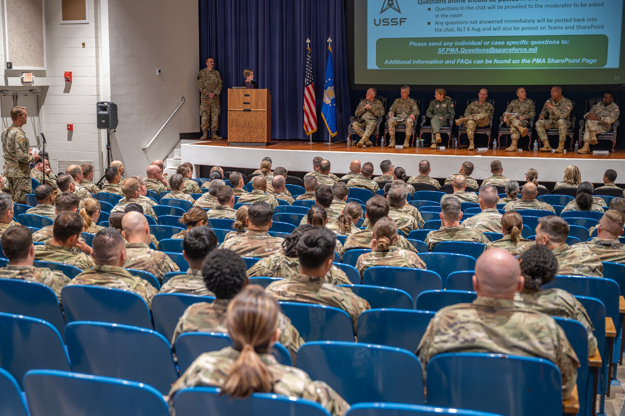People wearing military uniforms sitting in chairs on a stage overlooking a seated audience.