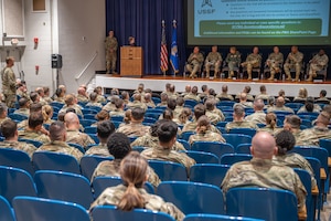 People wearing military uniforms sitting in chairs on a stage overlooking a seated audience.
