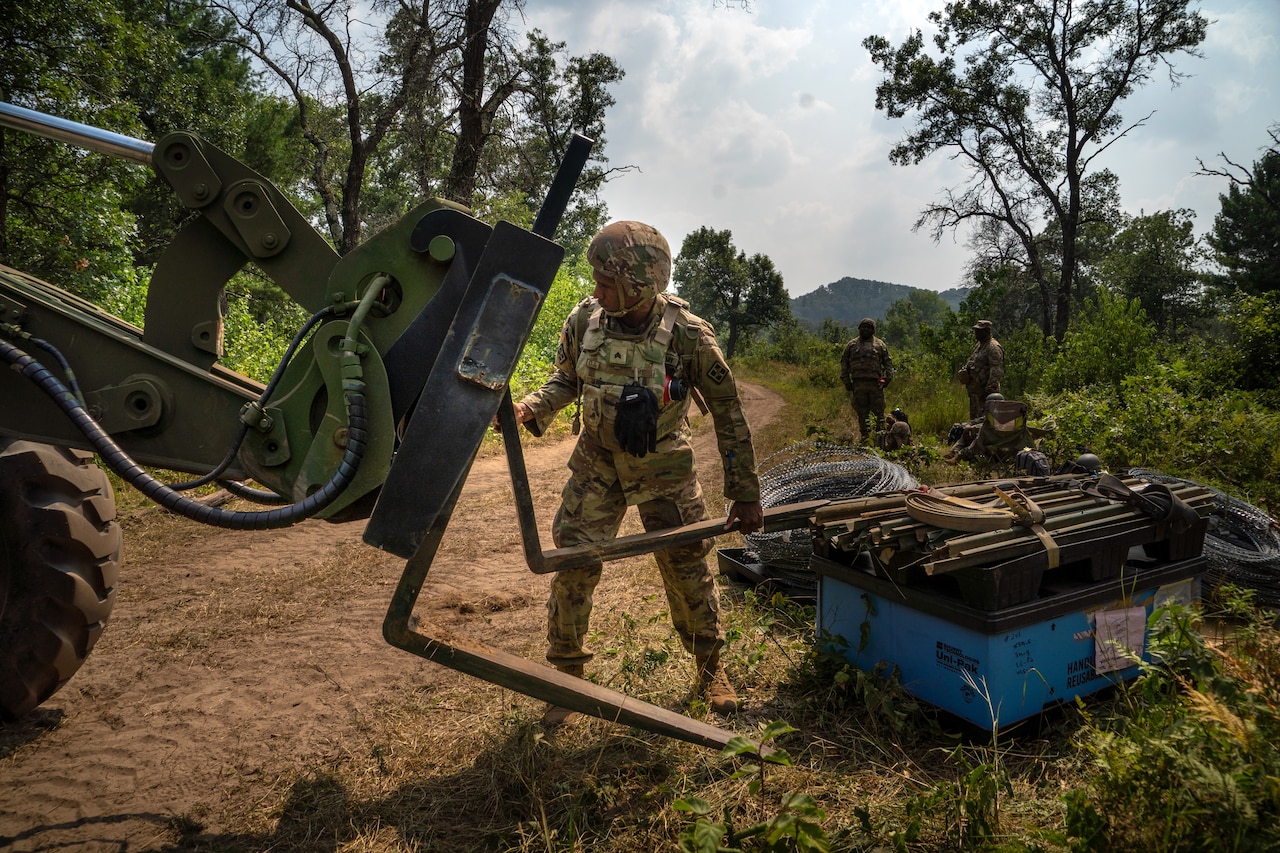 A soldier in a camouflage uniform moves a large steel forklift arm on a dirt road. There are several soldiers in camouflage uniforms in the background.