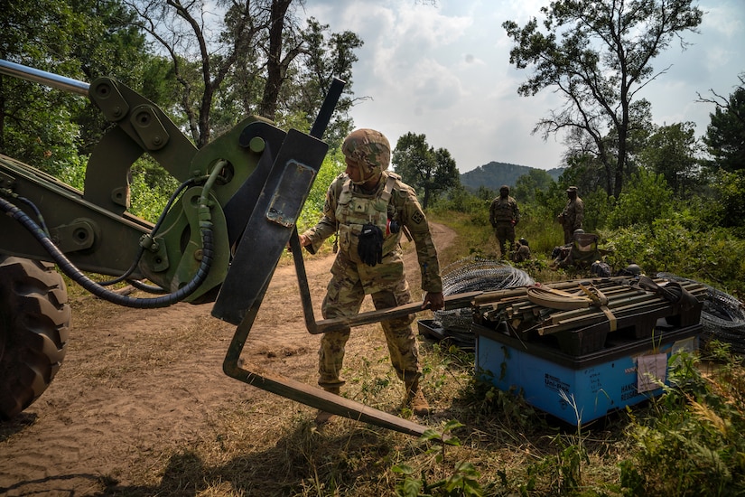 A soldier in a camouflage uniform moves a large steel forklift arm on a dirt road. There are several soldiers in camouflage uniforms in the background.