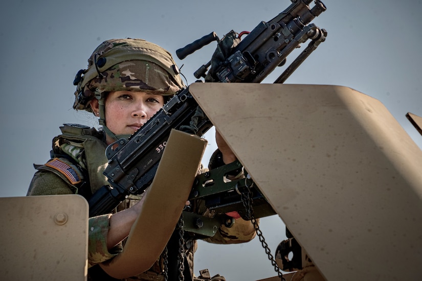 A soldier in a camouflage uniform and a helmet mans a large machine gun while looking at the camera.