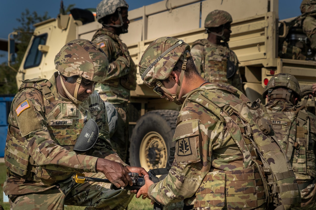 Two soldiers in camouflage uniforms assemble a machine gun. There are several soldiers in camouflage uniforms and a large military truck in the background.