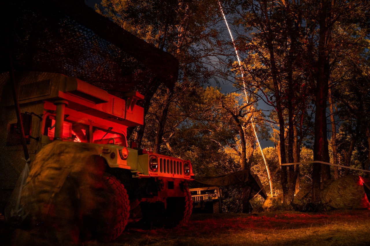 Two Humvees are parked in the foreground as a flash of light in the night from a projectile is seen in the background