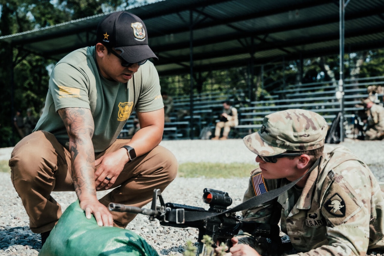 A man wearing sunglasses and a baseball cap kneels near a soldier wearing a camouflage uniform. The soldier is lying in the prone position with a rifle in his hands.