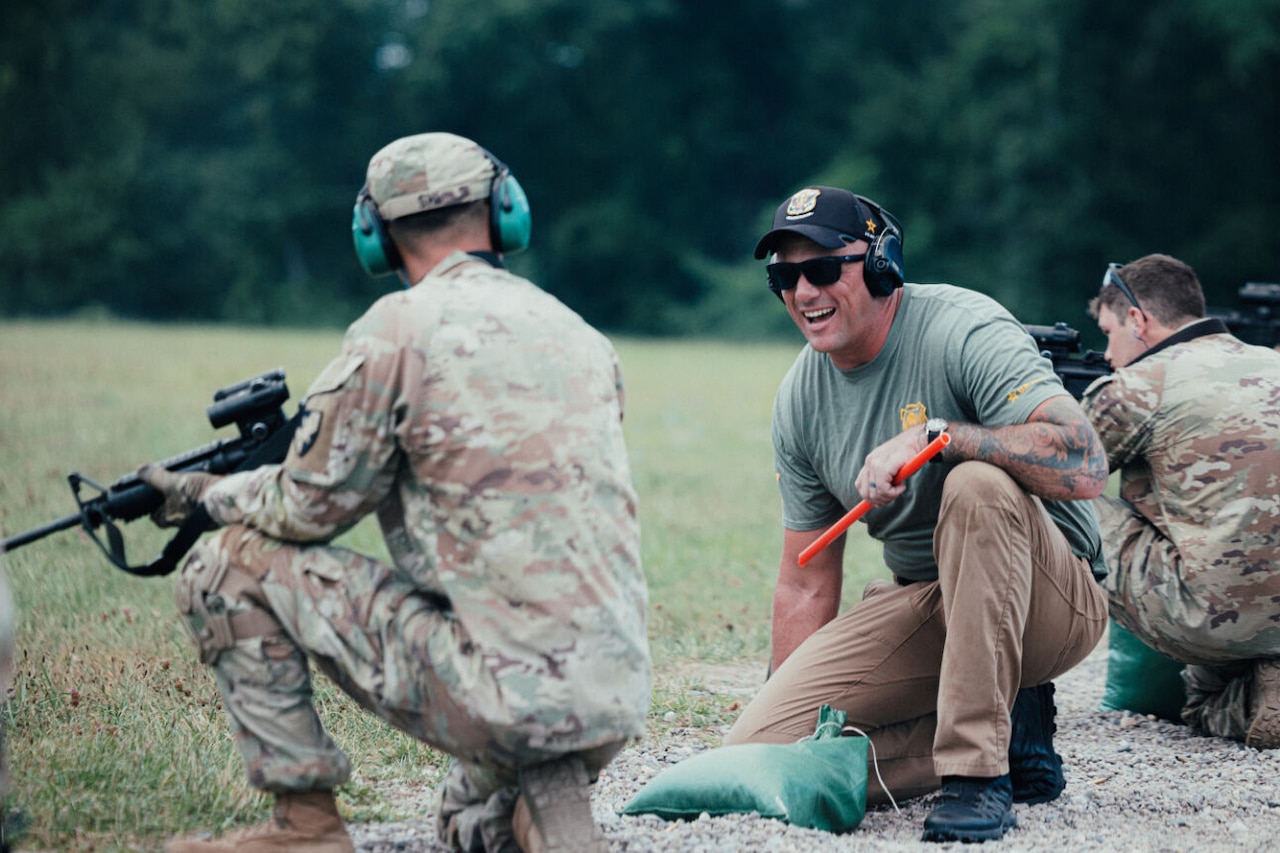 A man wearing sunglasses smiles and kneels next to a soldier in a camouflage uniform wearing hearing protection and holding a rifle. There are two soldiers in camouflage uniforms in the background aiming rifles.