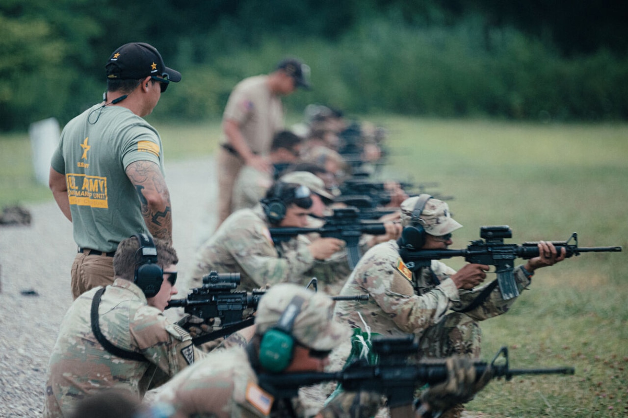 A man wearing a U.S. Army Marksmanship Unit T-shirt looks at soldiers in camouflage uniforms who are kneeling and aiming rifles at targets. There is a man in the background dressed in casual attire.