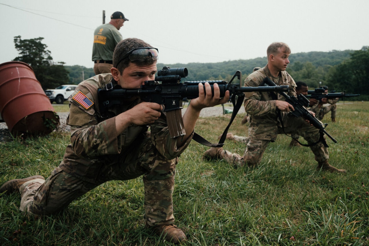 A soldier in a camouflage uniform is kneeling while aiming a rifle at a target. There are other soldiers kneeling in the background aiming or preparing to aim their rifles.