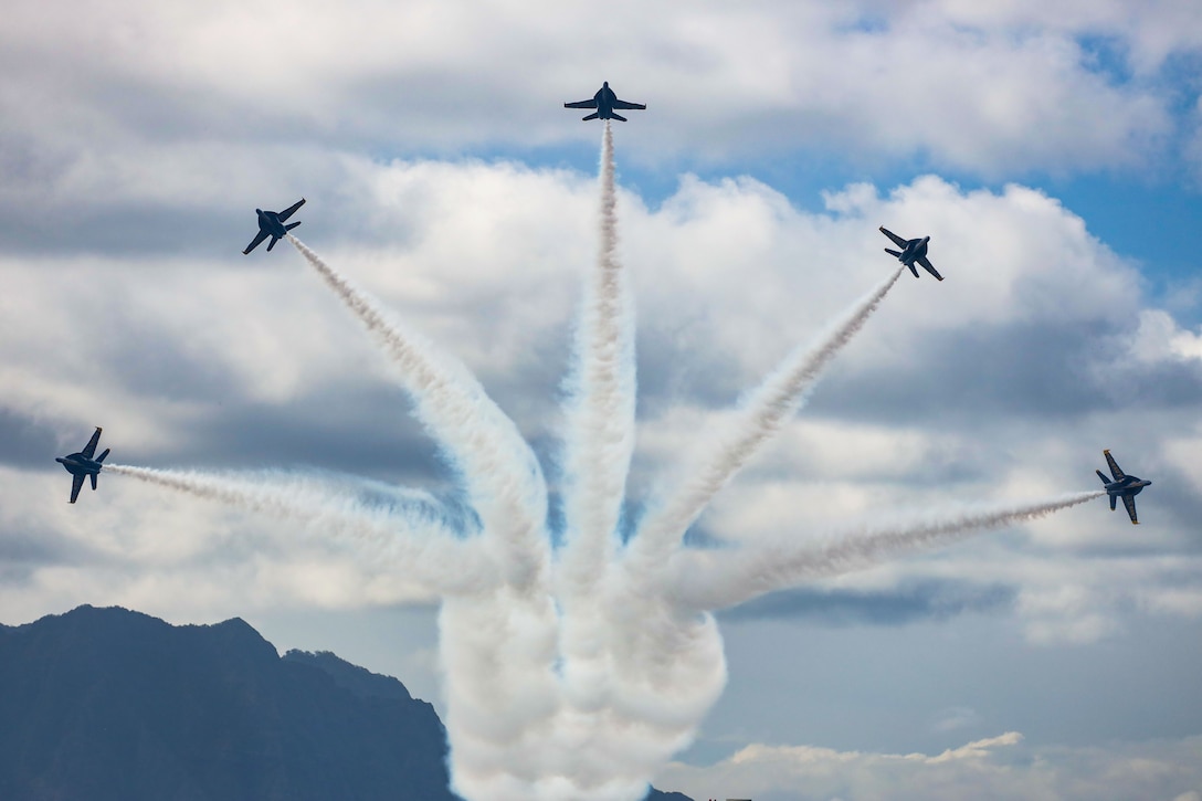 Five aircraft perform maneuvers in a blue sky leaving behind trails of smoke.