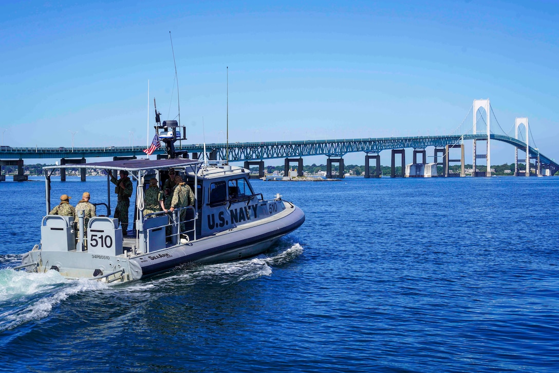 Sailors and college personnel ride in a small boat toward a bridge against a blue sky.