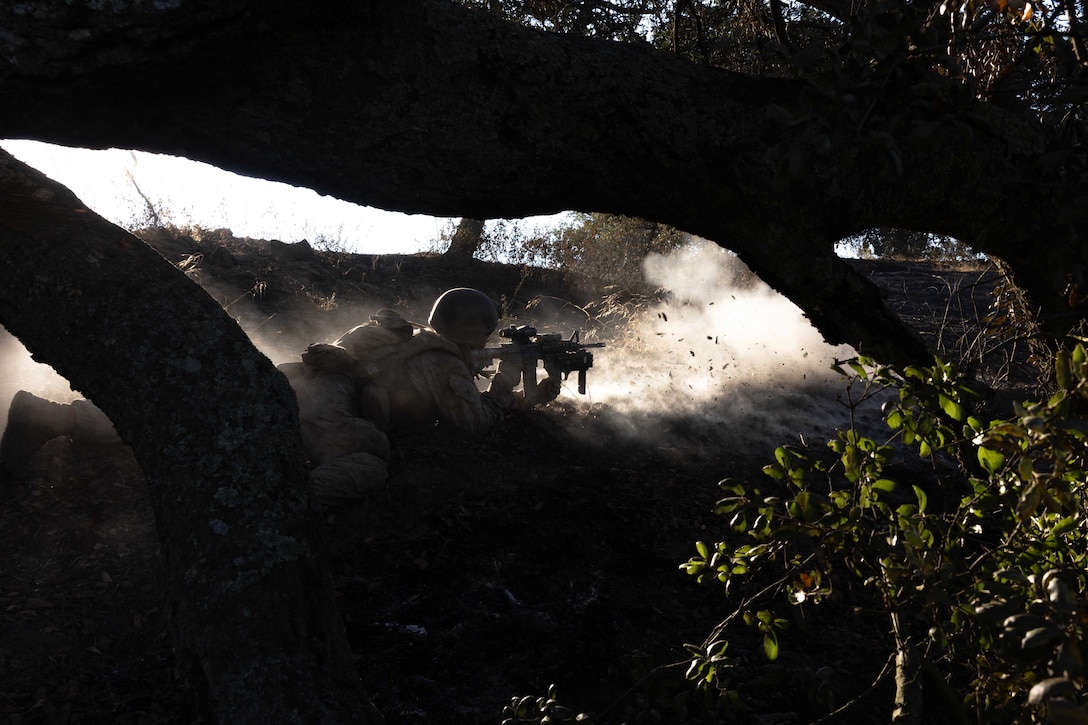 A Marine laying in a dark wooded area fires a weapon creating clouds of smoke.