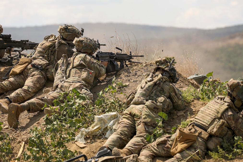 Soldiers in tactical gear lay on a dirt hill while firing weapons toward mountains in the distance.