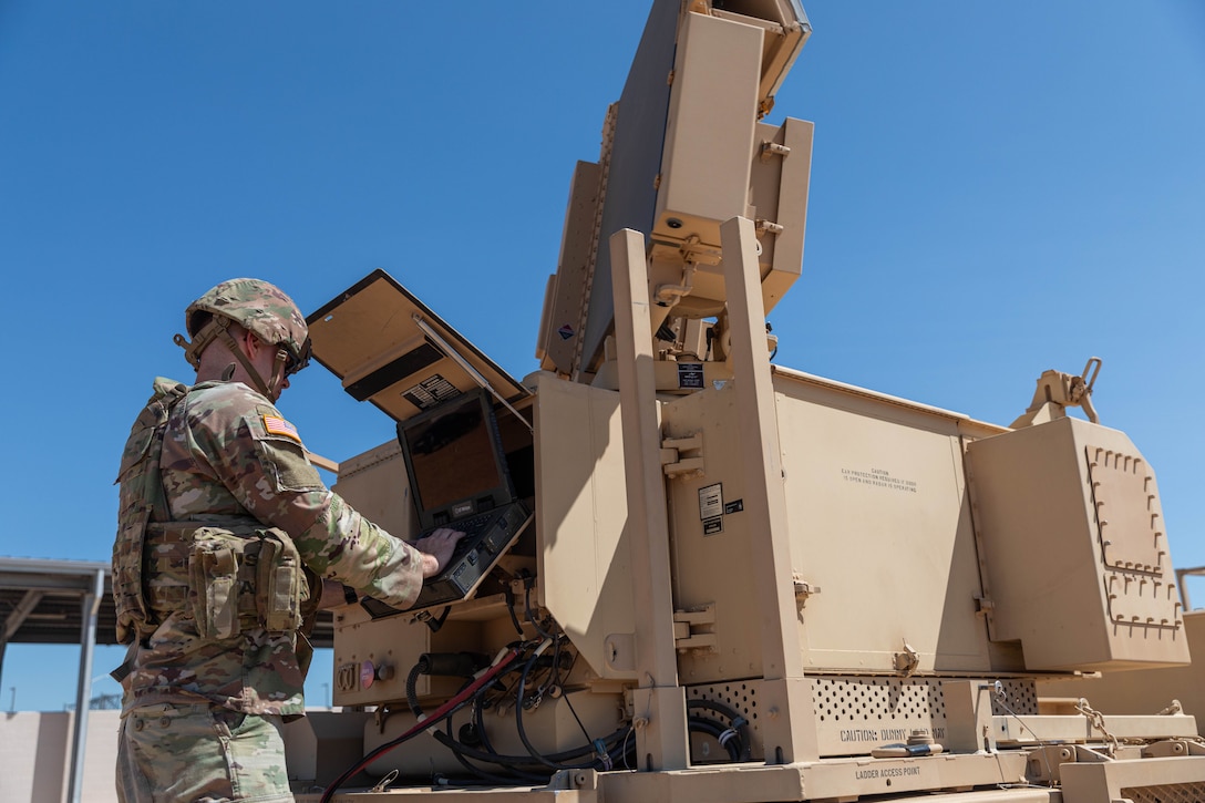 A soldier adjusts a large piece of tan machinery outside under a blue sky.