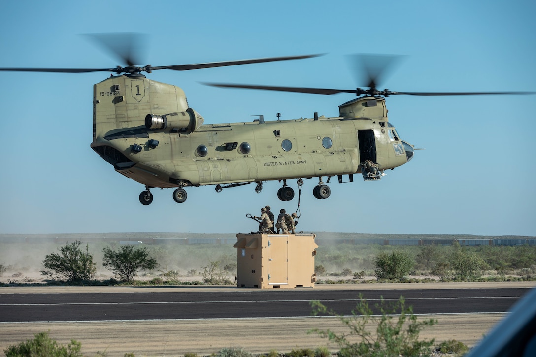 Soldiers standing in a container on a road hook the container to a helicopter in flight above them.