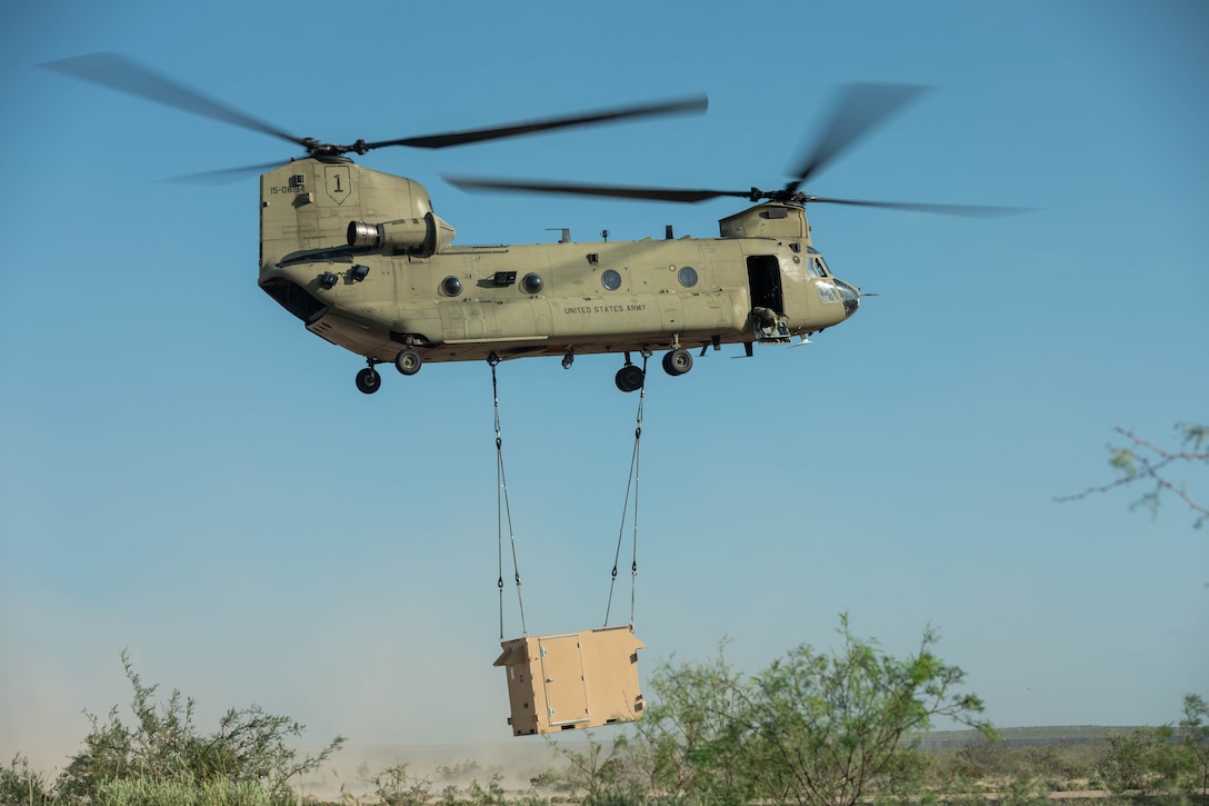 A helicopter carries a container as it flies above brush with a blue sky behind.
