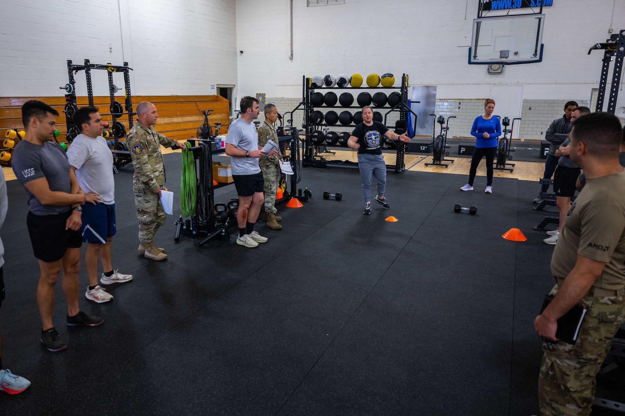 Military members in physical training gear listen to instructions for a physical challenge in a gym.