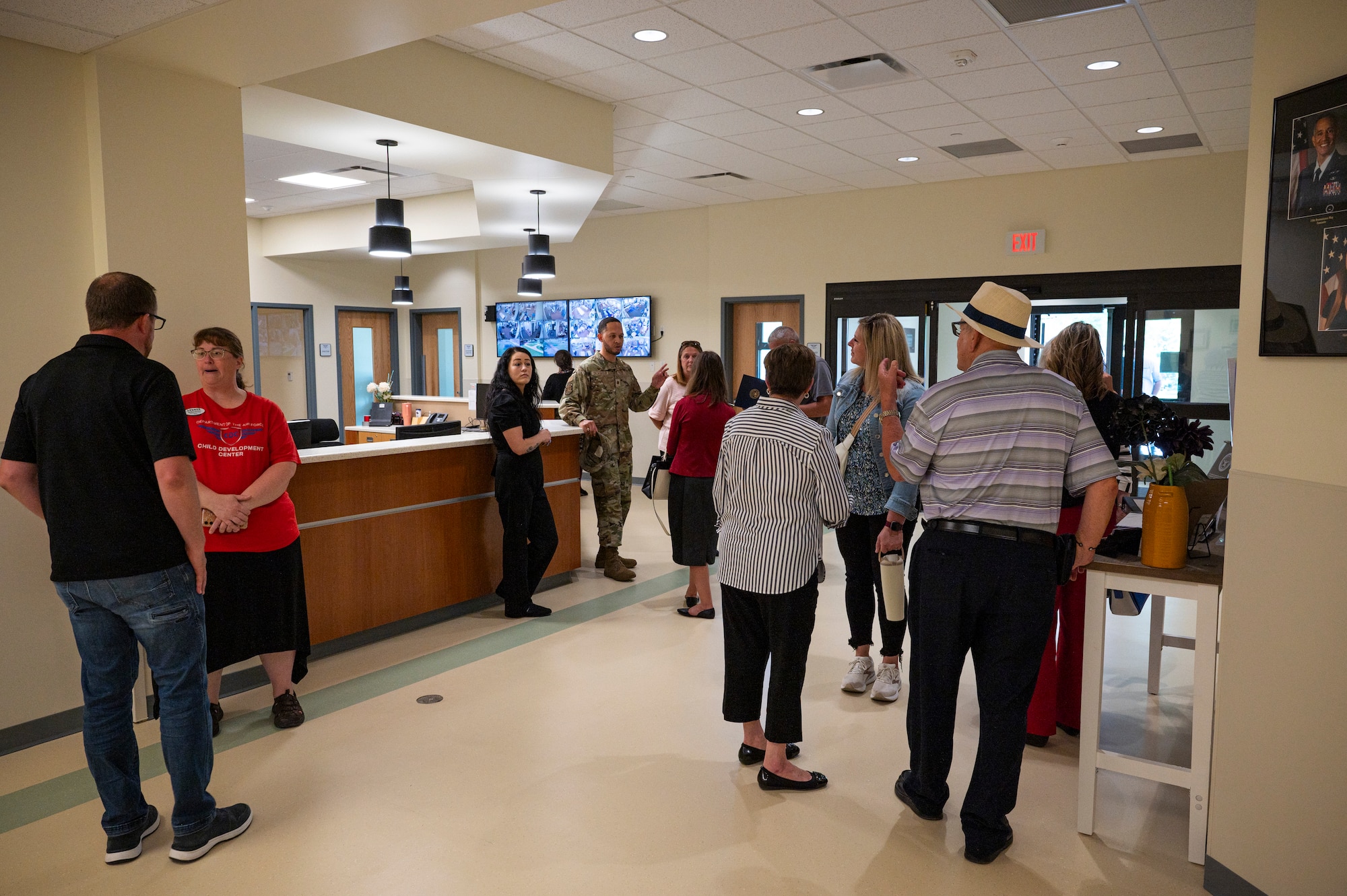 A large group of people walk around a room in a building, there are double doors leading into the room and a large wooden desk in the middle of the room.