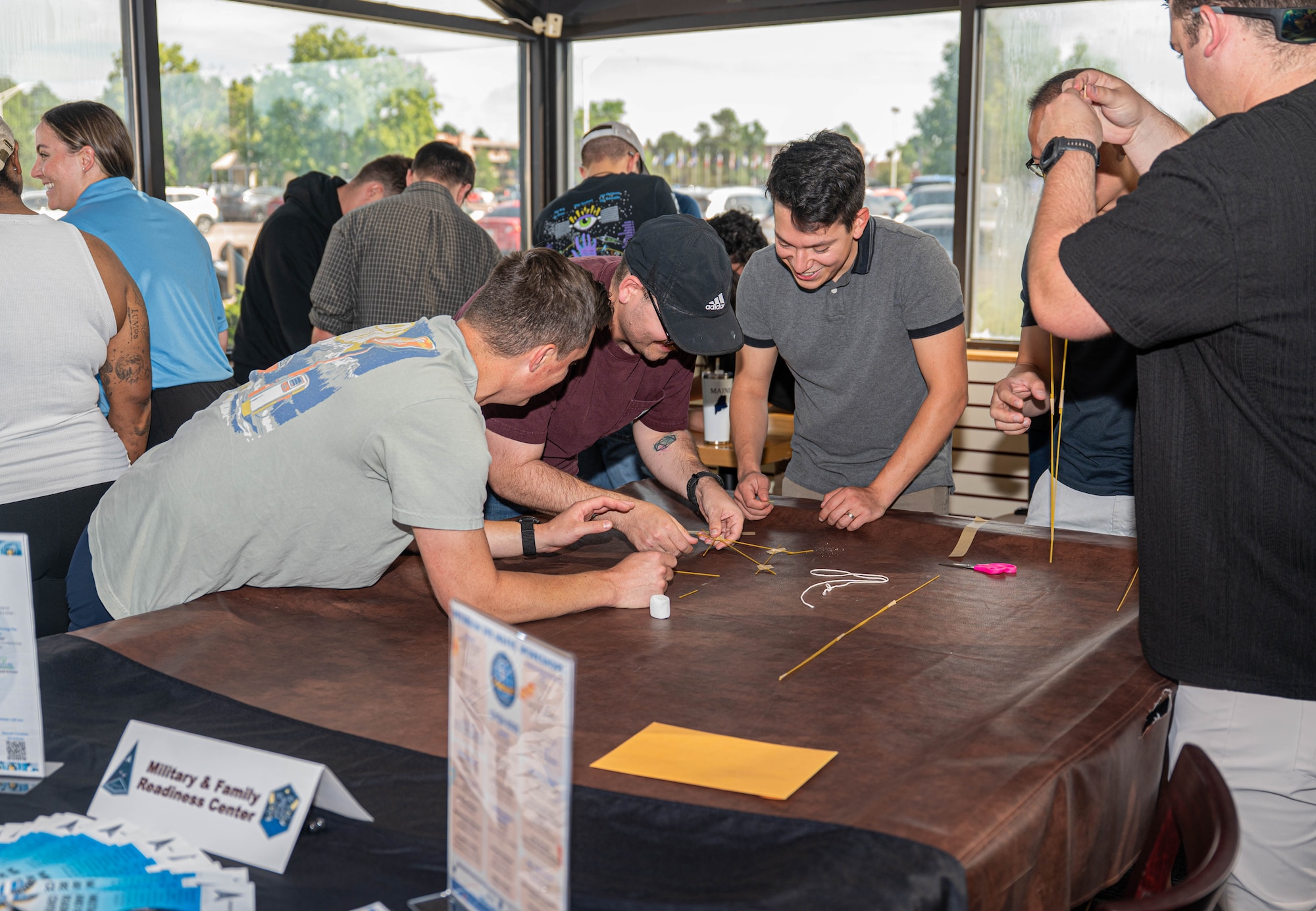 Members gathered together around a table to build a makeshift tower with unconventional materials.