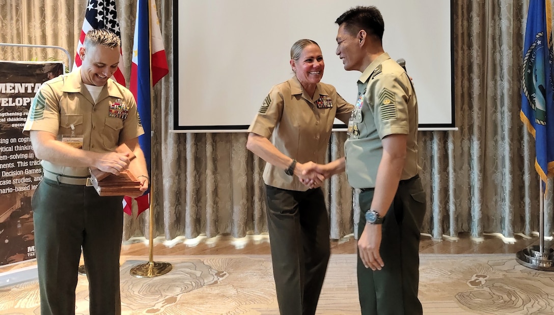 Sgt. Maj. Joy M. Kitashima, center, command senior enlisted leader, U.S. Marine Corps Forces, Pacific, thanks Fleet Command Master Sgt. Feliciano A. Lazo, right, sergeant major, Armed Forces of the Philippines, in front of group of senior enlisted leaders from several countries during the Pacific Amphibious Leaders Symposium, July 8, 2025, in Manila, Philippines.  As part of PALS, this was the first time senior enlisted leaders had a dedicated forum to come together to build relationships and discuss ways to better mentor, lead, and develop their enlisted forces.  Through discussions, engagements, and presentations, PALS enhances cooperation, tackles common threats, protects shared resources, and maintains sovereignty in the Indo-Pacific. This year’s symposium hosted senior leaders from 23 participating nations who are committed to a free and open Indo-Pacific, with the objective of strengthening and developing regional relationships. Master Gunnery Sgt. Jeremy M. Vought, left, senior enlisted advisor of communication strategy and operations, MARFORPAC, prepares a gift to be presented to Lazo. (Photo courtesy of the Philippine Marine Corps)