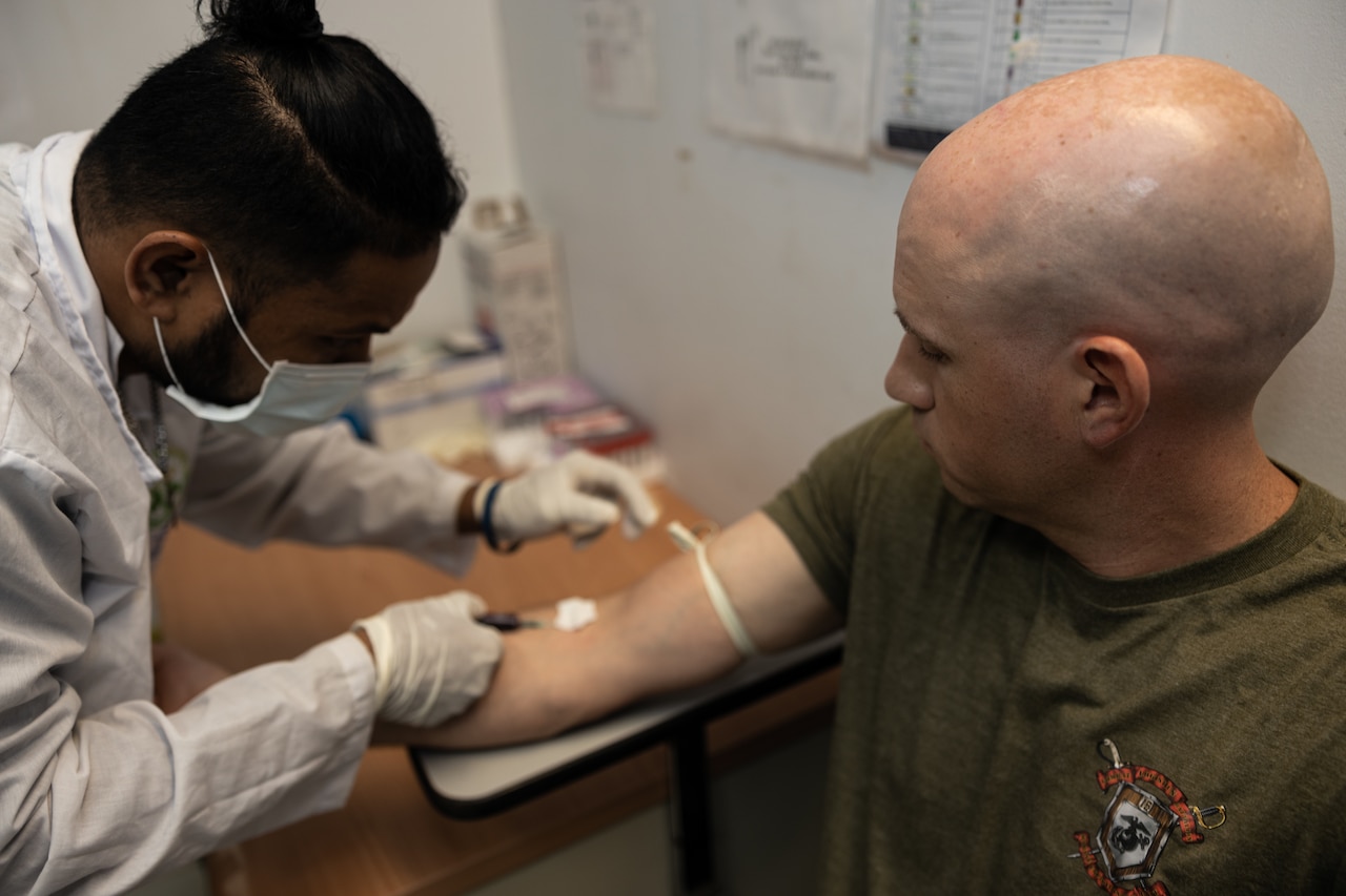 A man wearing a lab coat, gloves and a face mask, places a needle into the arm of another man wearing a green T-shirt with a crest printed on it to draw blood.