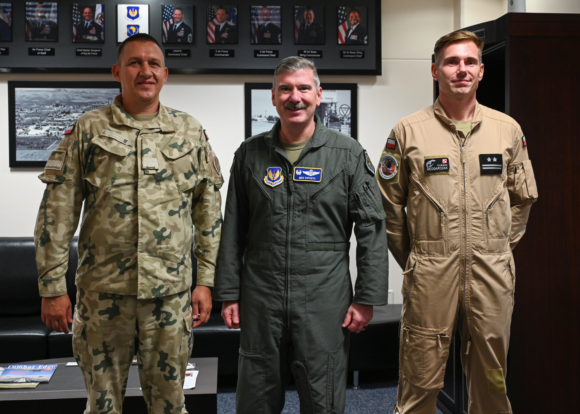 three military members, two polish, one american, posing and smiling in front of a wall with paintings