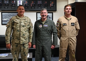 three military members, two polish, one american, posing and smiling in front of a wall with paintings