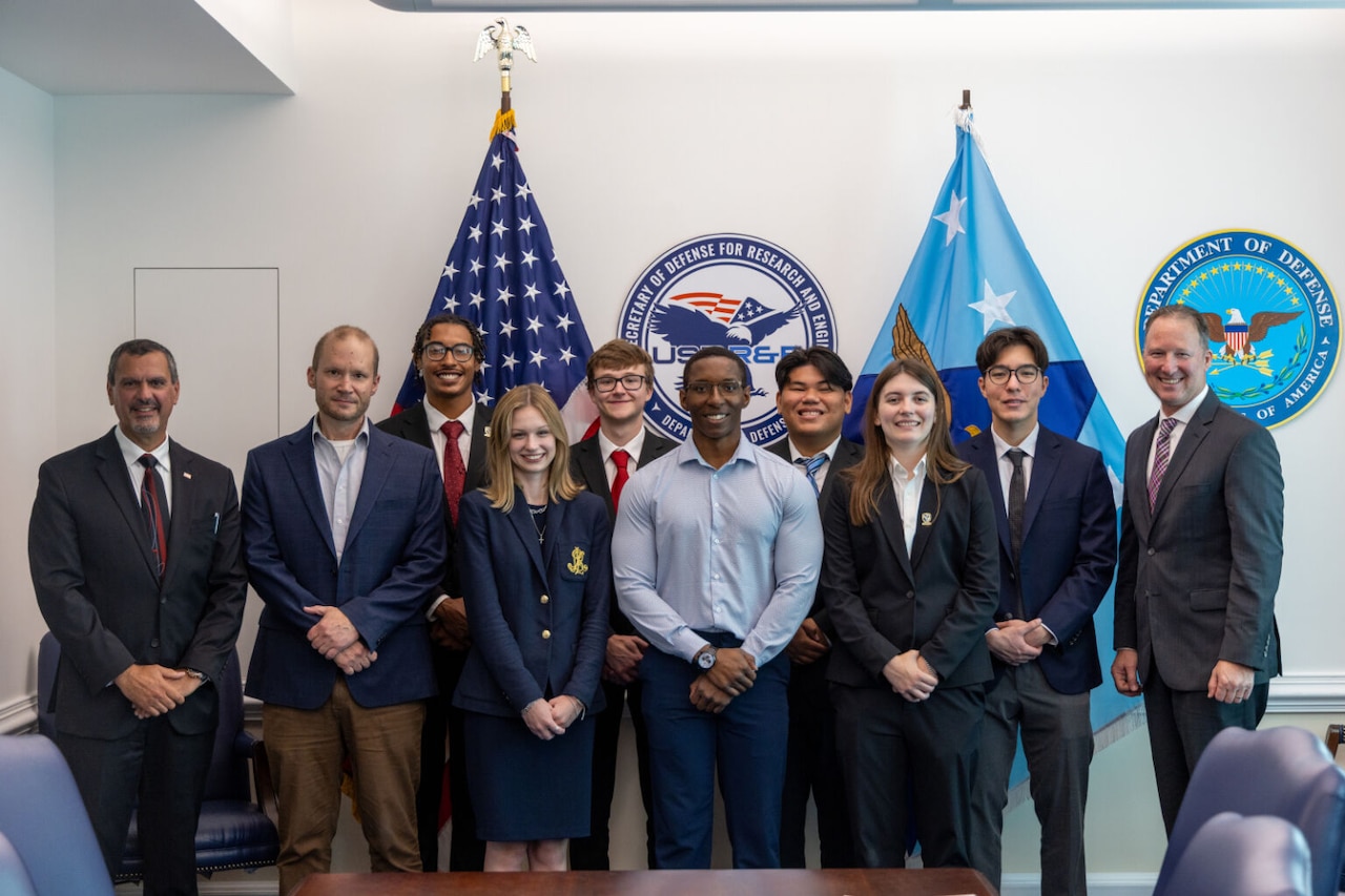 Several men and women in business attire pose for a photo with the American and Defense Department flags in the background.