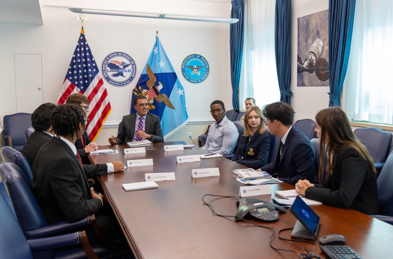 A man in a business suit sits at a long table while speaking to several young adults in business attire who are gathered at the table. An American flag and a DOD flag are displayed in the background.