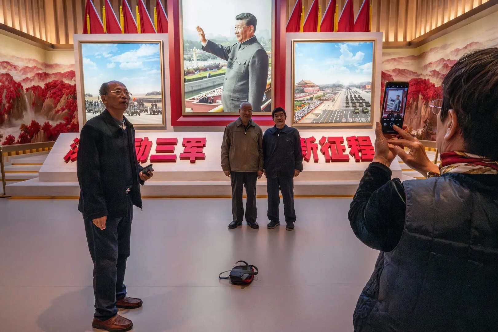 Four adults pose inside a museum or exhibition space in front of large framed images of Xi Jinping and a military parade. Two men stand together in the center for a photo, while a woman on the right takes their picture with a smartphone. Another man stands to the left holding a phone. Red Chinese characters and decorative murals with mountains and waterfalls surround the display.