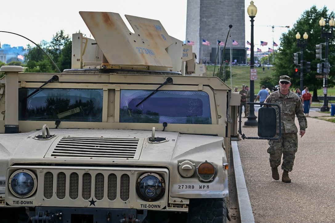 A soldier walks on a sidewalk near a parked Humvee during daytime, with other soldiers and people, American flags, and a monument in the background.