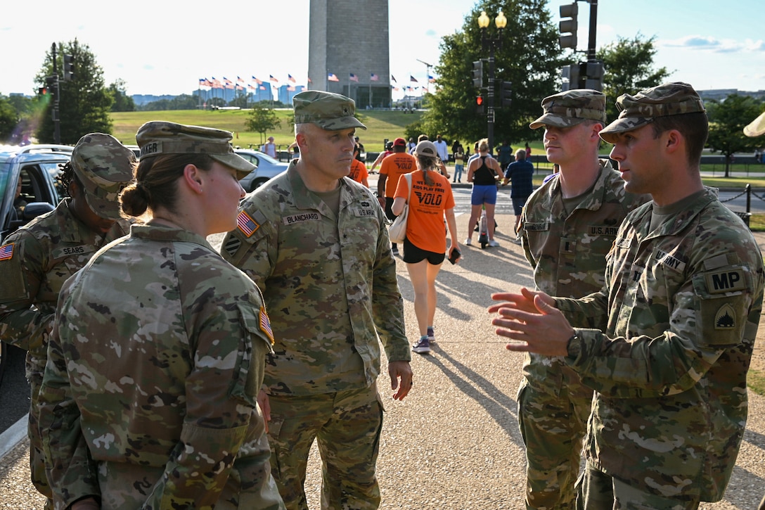 Five soldiers stand in a circle and talk on a sidewalk during daytime, with vehicles, people, a grassy field, American flags and a monument in the background.