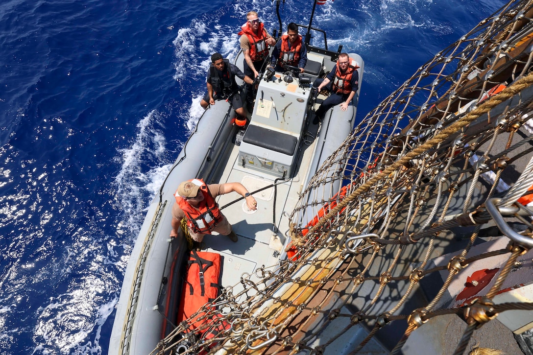 Five sailors in a small boat pull up next to netting attached at a ship at sea during the day.
