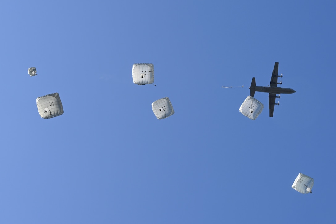 Seven soldiers descend with parachutes under an aircraft in a blue sky as seen from below.