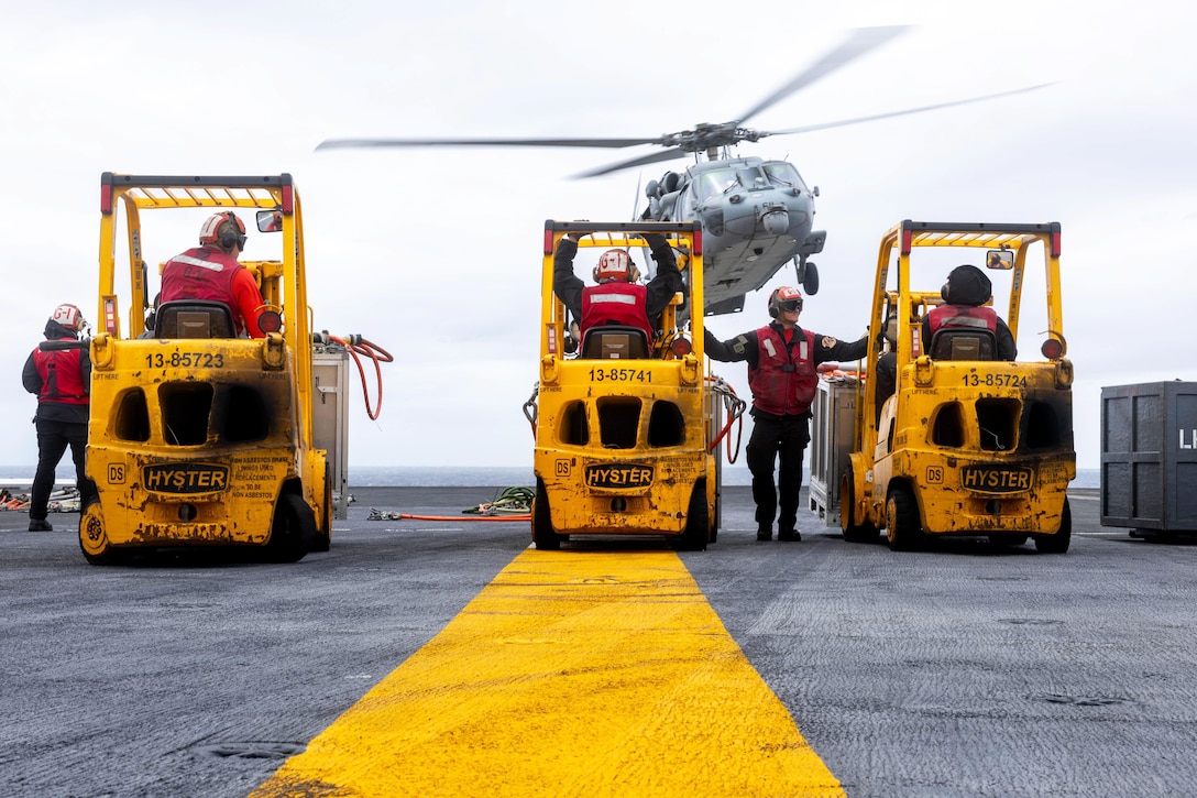Three sailors in forklifts and two standing nearby watch as a helicopter takes off from a ship at sea on a gloomy day.