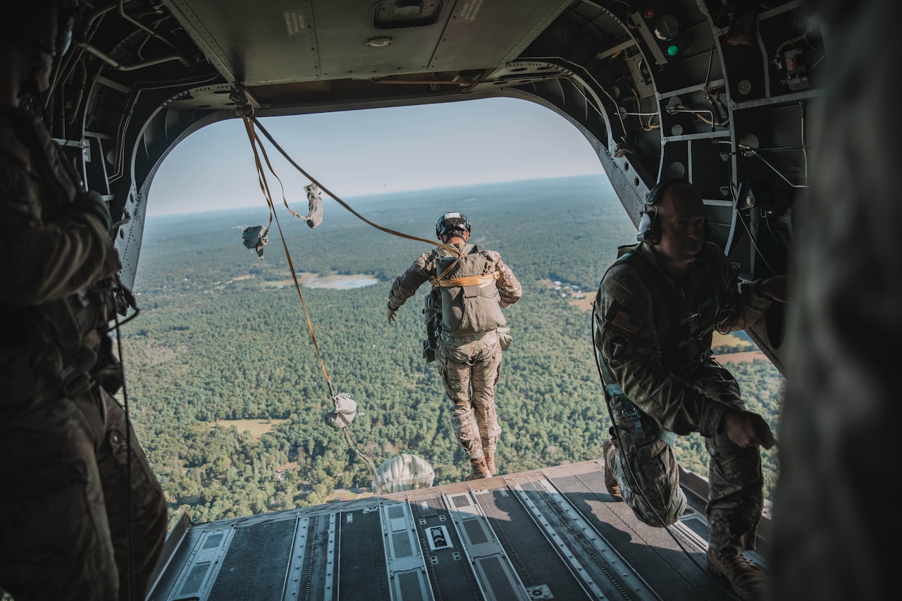 A soldier in a parachute jumps out of an aircraft in flight over a forest on a sunny day.