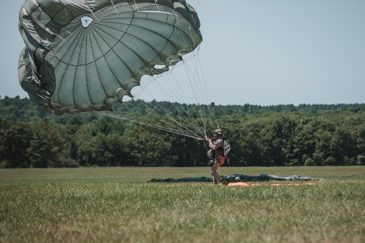 A soldier in a parachute lands in a field with trees in the background on a sunny day.
