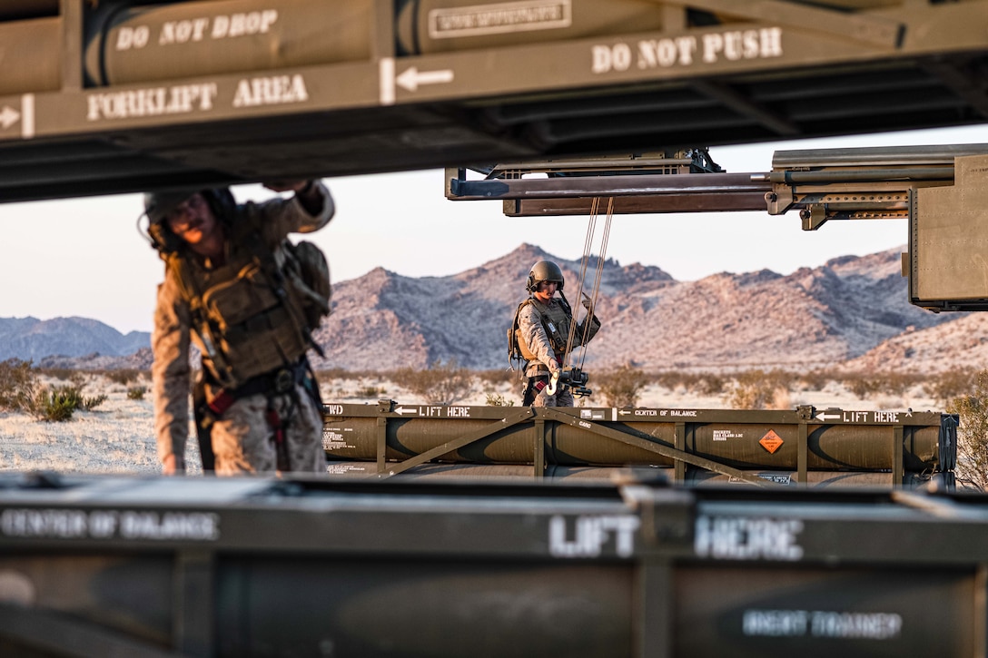 A close-up of two Marines in tactical gear loading a rocket launcher in a mountainous area.
