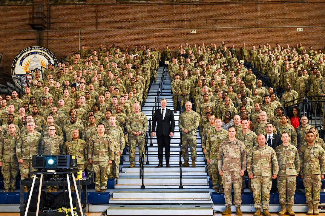 A person wearing a business suit stands with a large group of soldiers and airmen indoors, with a wall in the background.