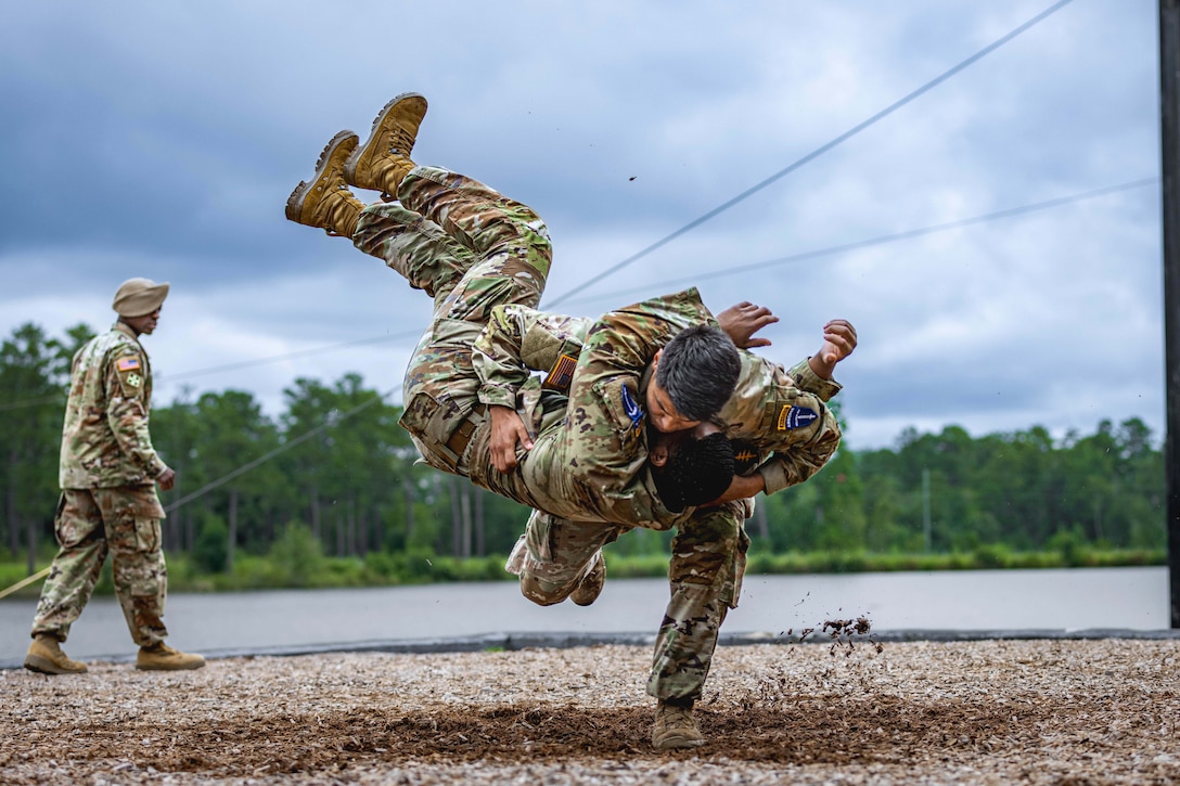 A soldier takes down a fellow soldier on woodchips as another watches from the background against a wooded area.