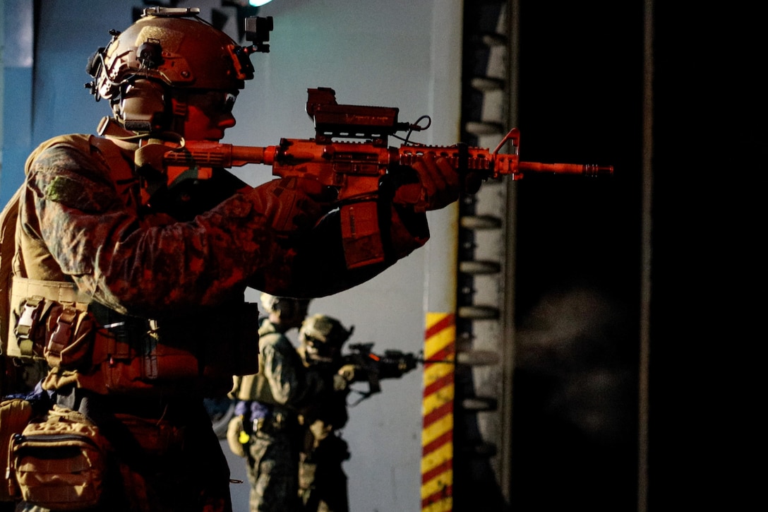 A close-up of a Marine in tactical gear aiming a weapon preparing to enter a dark area as fellow Marines do the same in the background.