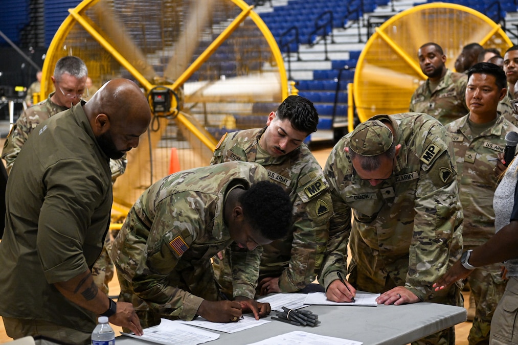 A marshal helps National Guardsmen sign paperwork at a table in a gymnasium as two large fans blow in the background.