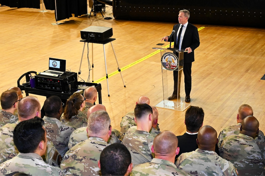 A person wearing a business suit stands and speaks at a lectern to a group of guardsmen indoors.
