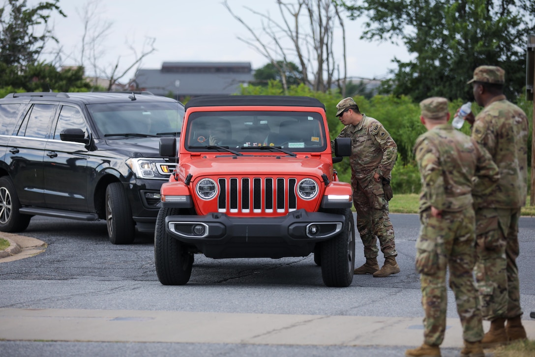 A soldier talks to the driver of a vehicle stopped on a driveway-like road, as two other soldiers look on; another vehicle is behind the stopped vehicle.